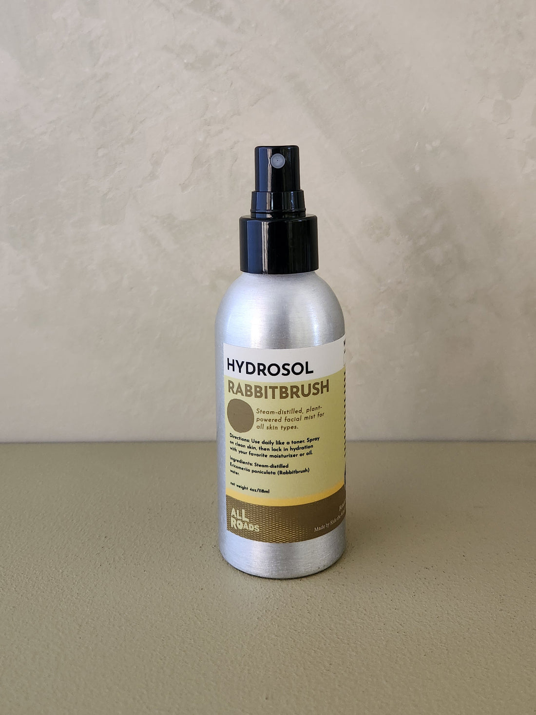 Hydrosol Rabbitbrush room spray in a silver aluminum bottle with a black spray nozzle and a beige and brown label, set against a textured beige background.