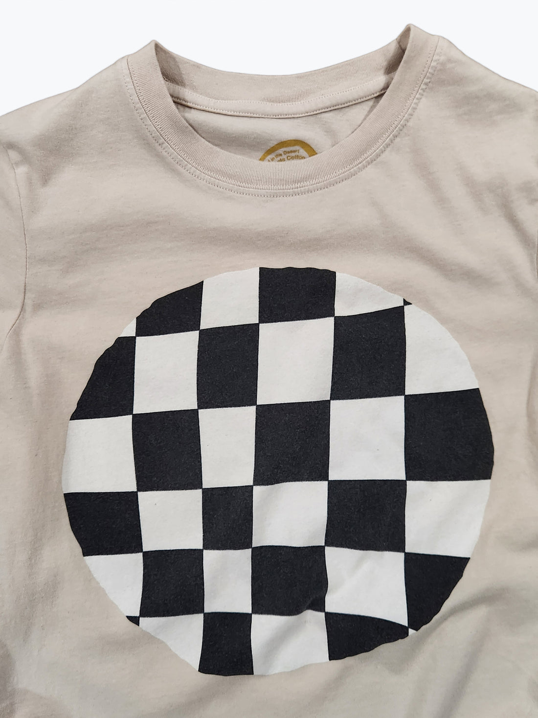 A close-up of a beige crewneck t-shirt featuring a large circular black and white checkerboard graphic printed on the front.
