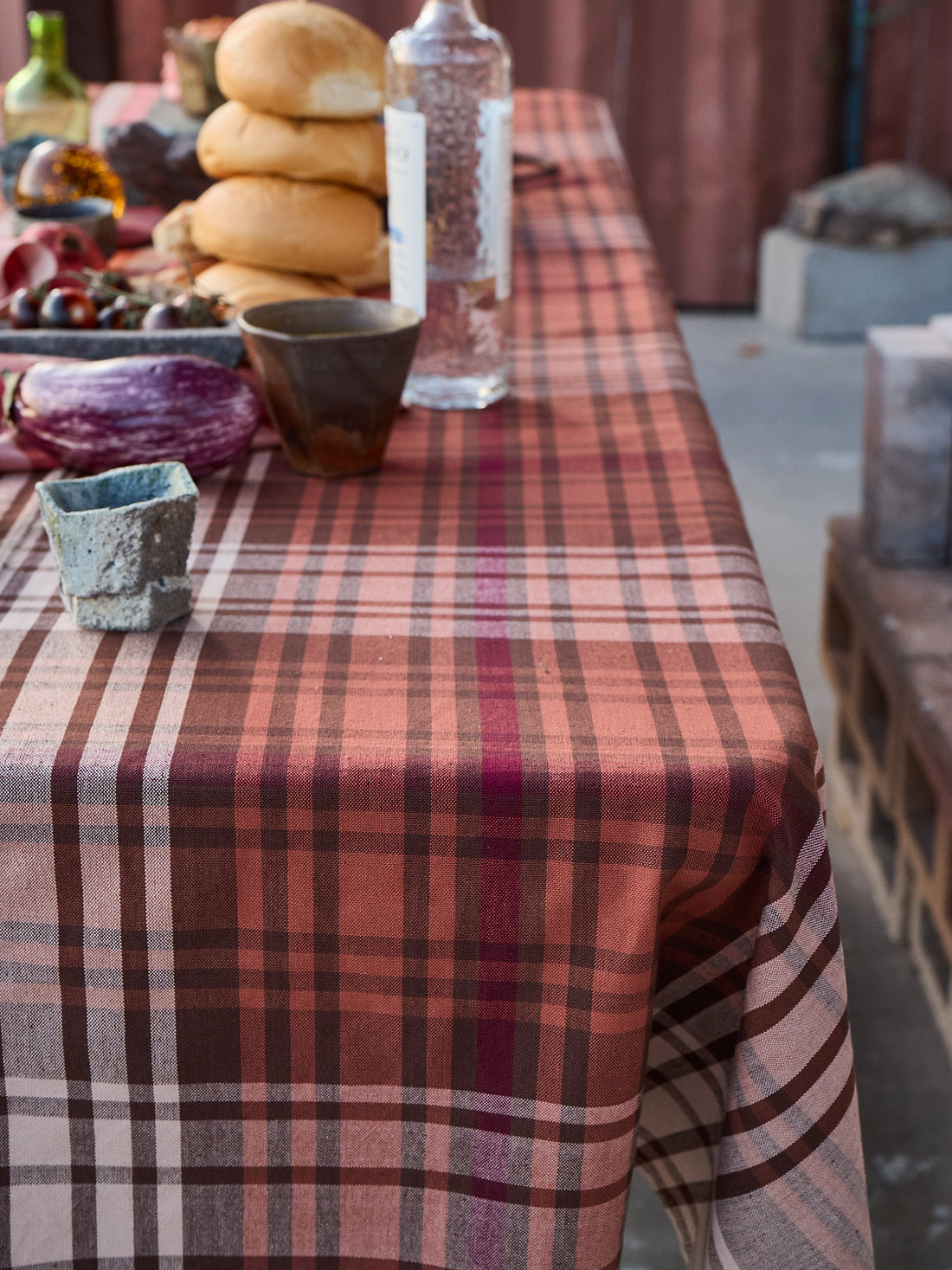 Table with a plaid tablecloth, various items including bread and bottles, on an outdoor setting.