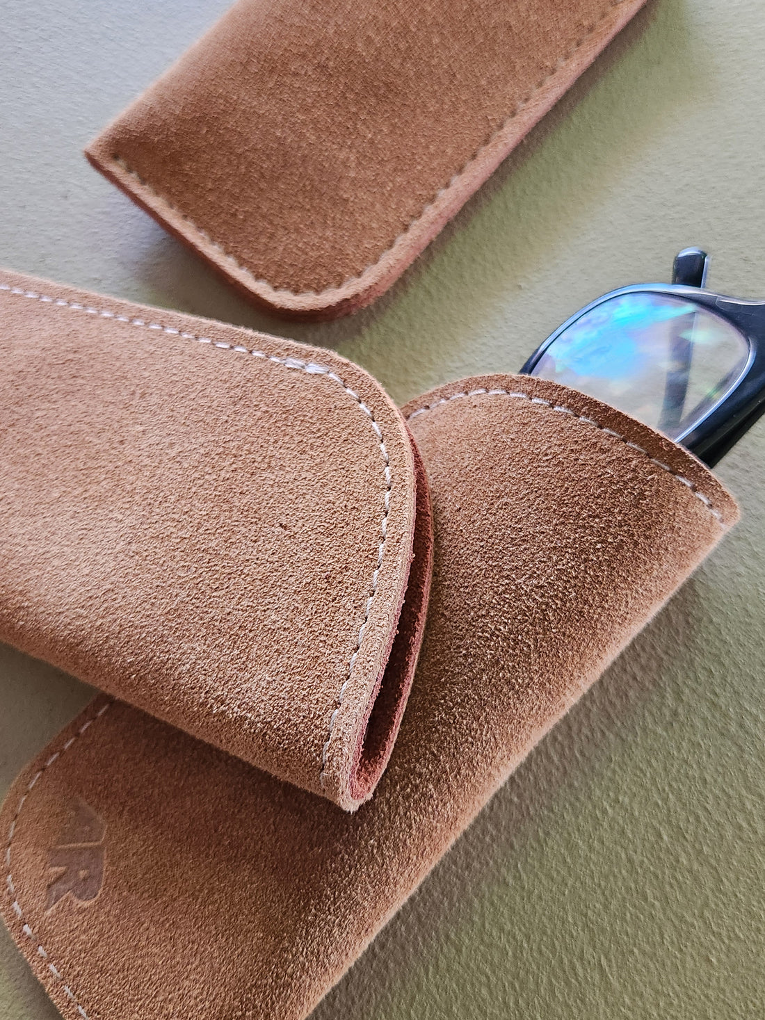 Brown suede eyeglass case on a light green surface with glasses partially visible.