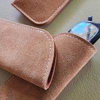 Brown suede eyeglass case on a light green surface with glasses partially visible.