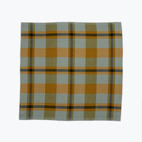 A square plaid napkin in muted sage green, mustard yellow, and dark brown stripes, displayed flat against a clean white background.