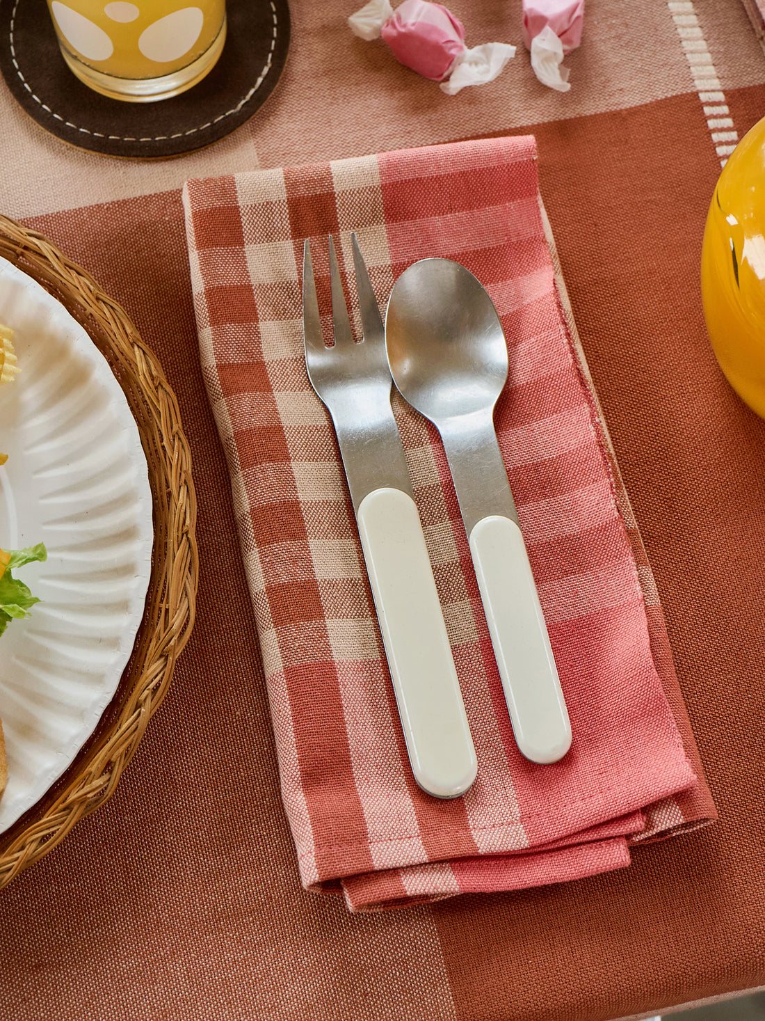 Silver fork and spoon on a pink checkered napkin with a plate and cup in the background.
