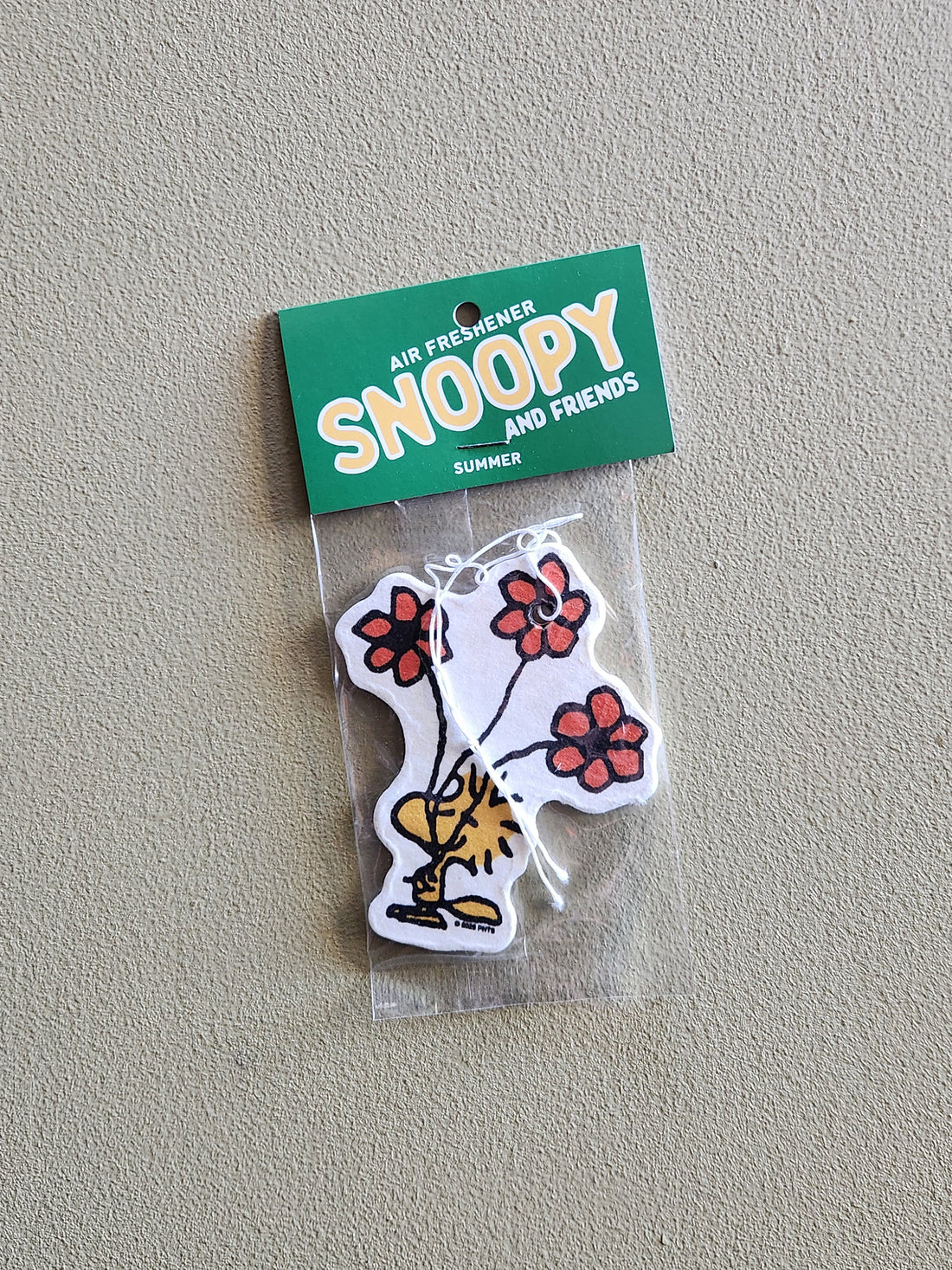 A Snoopy and Friends air freshener sticker in its clear plastic packaging with a green header card, placed on a textured beige wall. The sticker features a white dog body with red flowers and a yellow bird on it.
