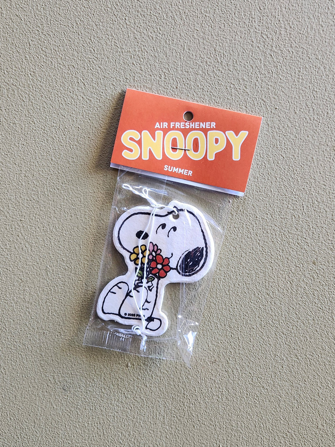 A Snoopy air freshener shaped like the character holding red and yellow flowers, sealed in a clear plastic bag with an orange header card that reads &