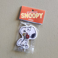 A Snoopy air freshener shaped like the character holding red and yellow flowers, sealed in a clear plastic bag with an orange header card that reads 'AIR FRESHENER SNOOPY SUMMER' on a textured beige background.