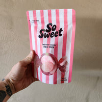 Hand holding a pink and white striped bag labeled 'So Sweet' against a beige background
