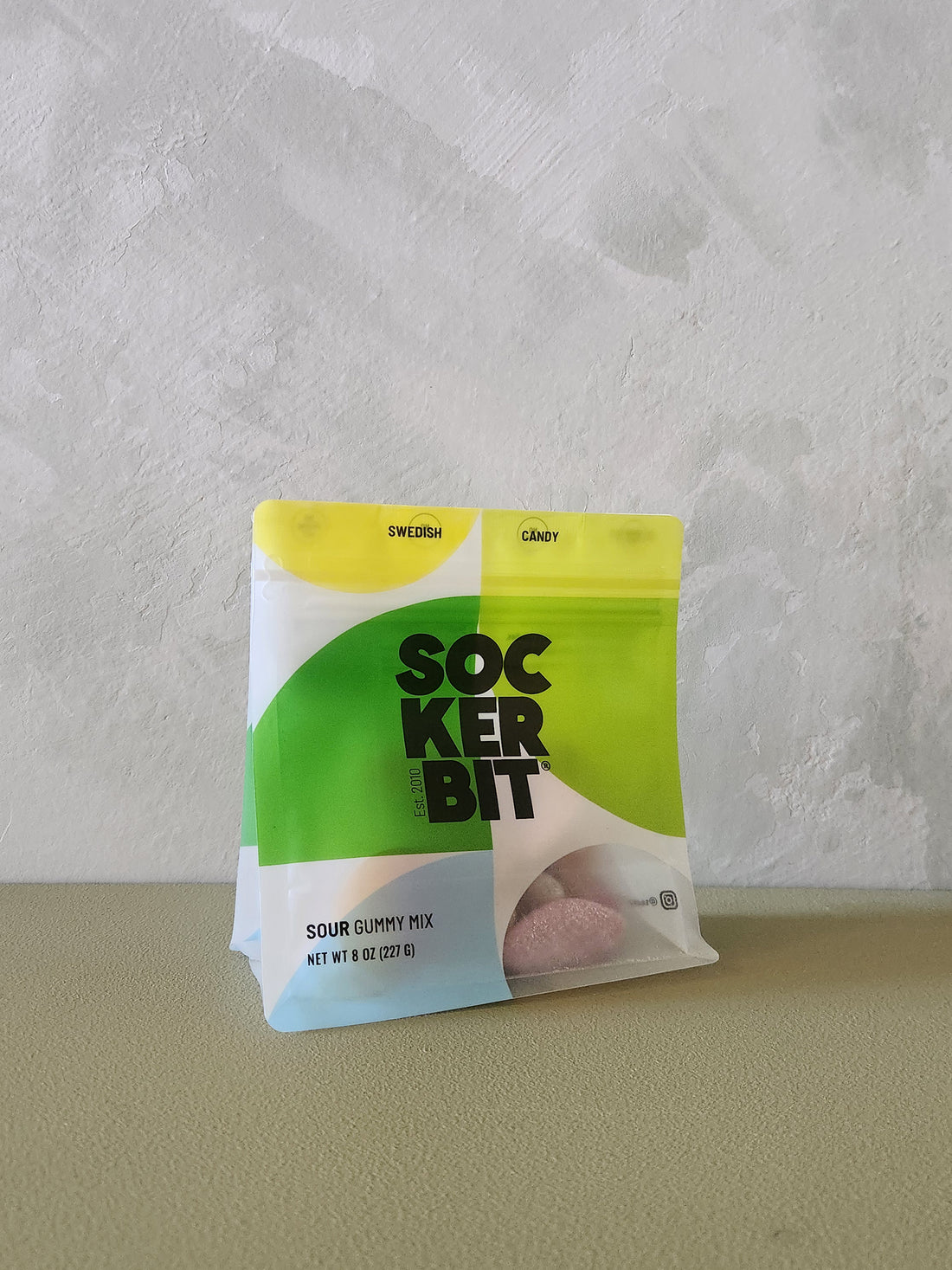 A bag of SockerBit Sour Gummy Mix, featuring a bright green and white packaging design with a yellow top section, standing on a textured beige surface against a light gray wall.