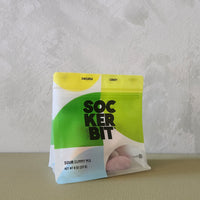 A bag of SockerBit Sour Gummy Mix, featuring a bright green and white packaging design with a yellow top section, standing on a textured beige surface against a light gray wall.
