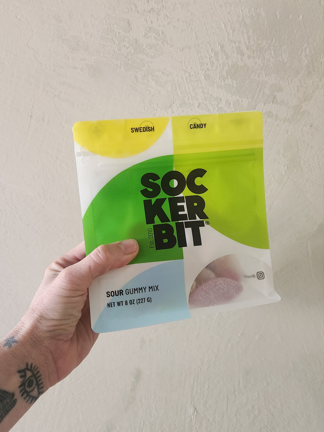 A hand holding a bag of Sockerbit Swedish Candy Sour Gummy Mix against a textured off-white wall.