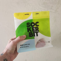 A hand holding a bag of Sockerbit Swedish Candy Sour Gummy Mix against a textured off-white wall.