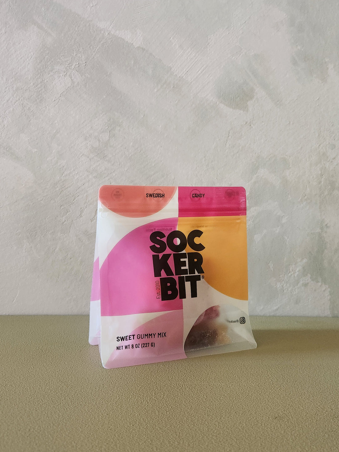 A bag of Sockerbit Sweet Gummy Mix standing upright on a beige surface against a textured off-white wall. The packaging features a bold, modern design with pink, orange, and yellow geometric shapes and black text.