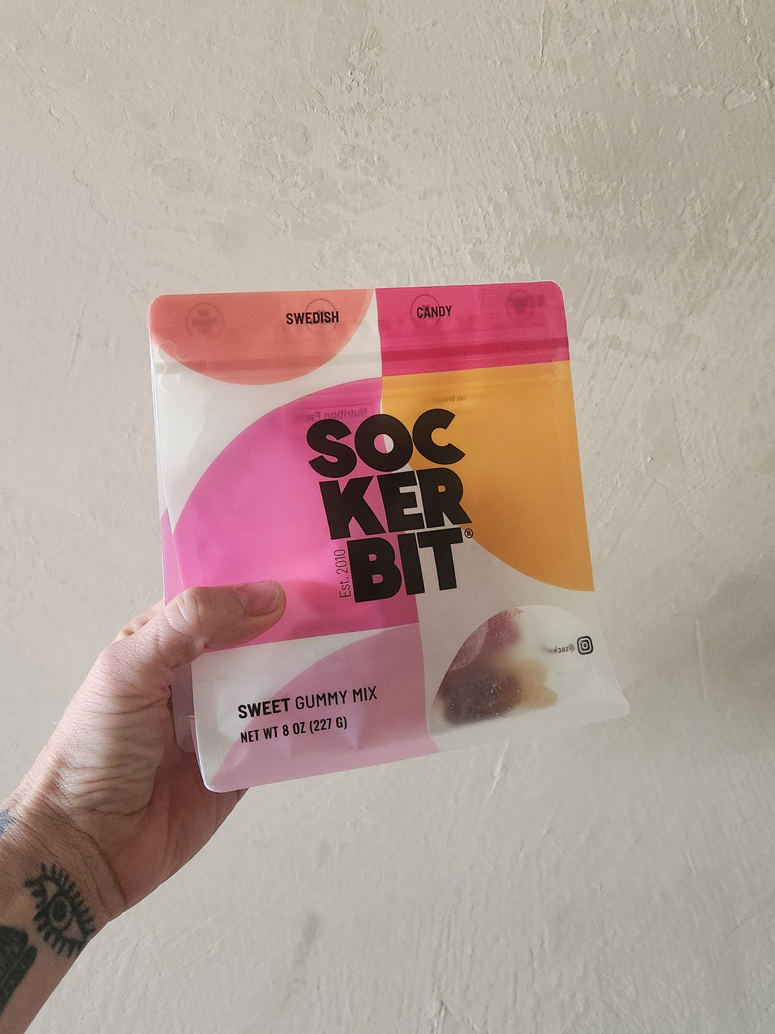 A hand with a tattoo holds a package of SockerBit Swedish Candy Sweet Gummy Mix against a textured off-white wall.