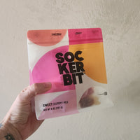 A hand with a tattoo holds a package of SockerBit Swedish Candy Sweet Gummy Mix against a textured off-white wall.