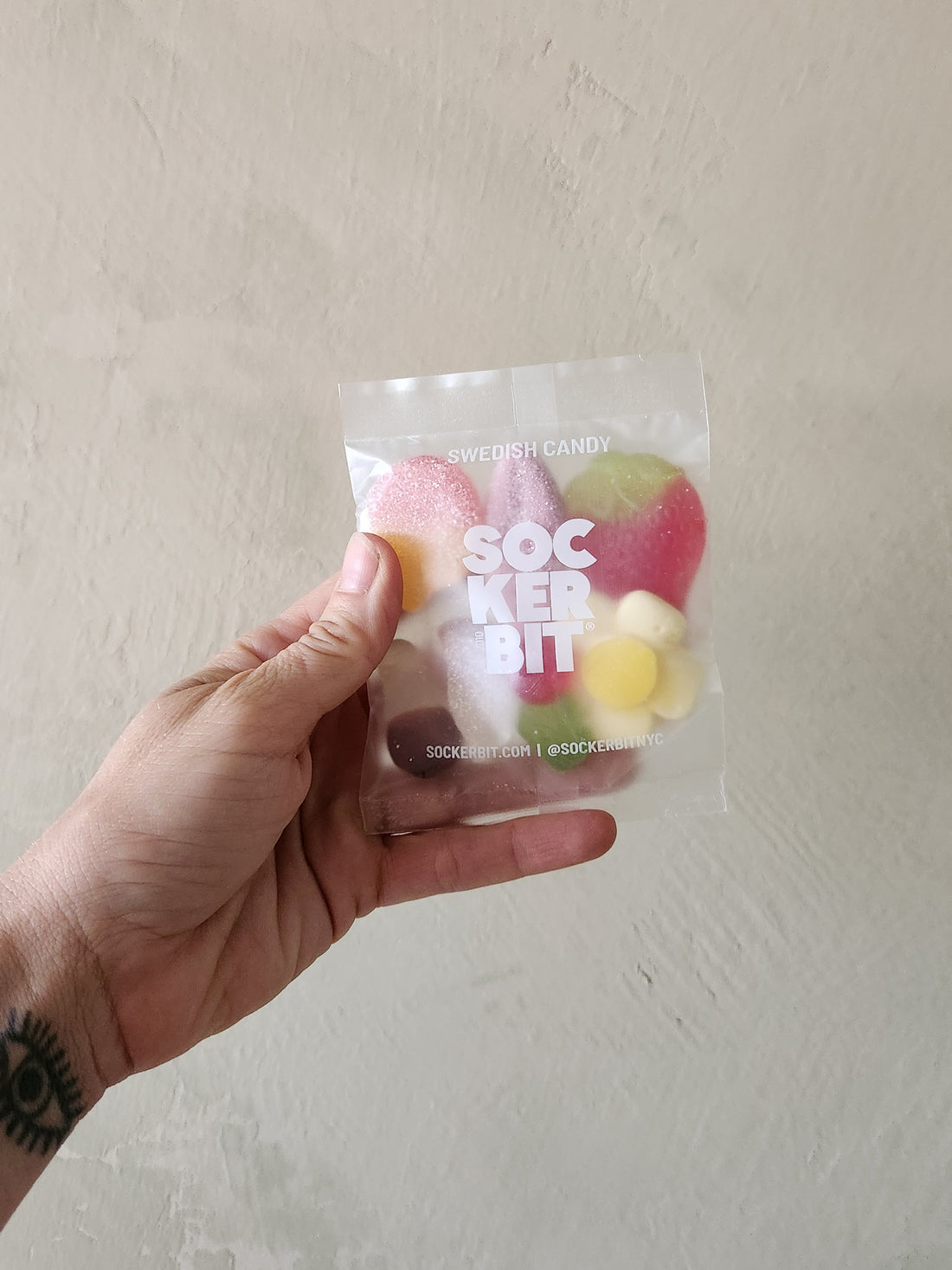 A hand holding a clear plastic bag of Swedish candy SOCKER BIT against a textured beige wall.