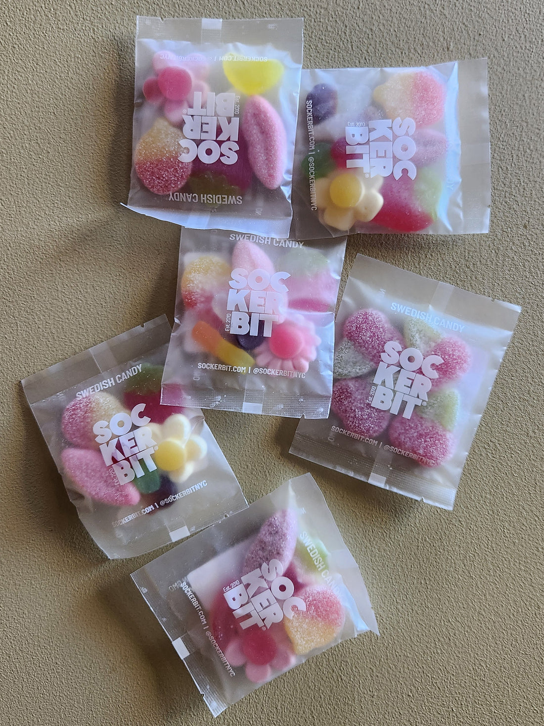Six clear plastic bags of Sockerbit  sour candy arranged on a textured beige surface. The candy inside is various colors including pink, yellow, and green, and the packaging features the Sockerbit logo.