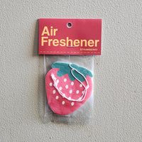 A strawberry-shaped air freshener in a clear plastic bag with a red header card that reads 'Air Freshener' and 'STRAWBERRY' in yellow text, photographed against a textured light gray wall.