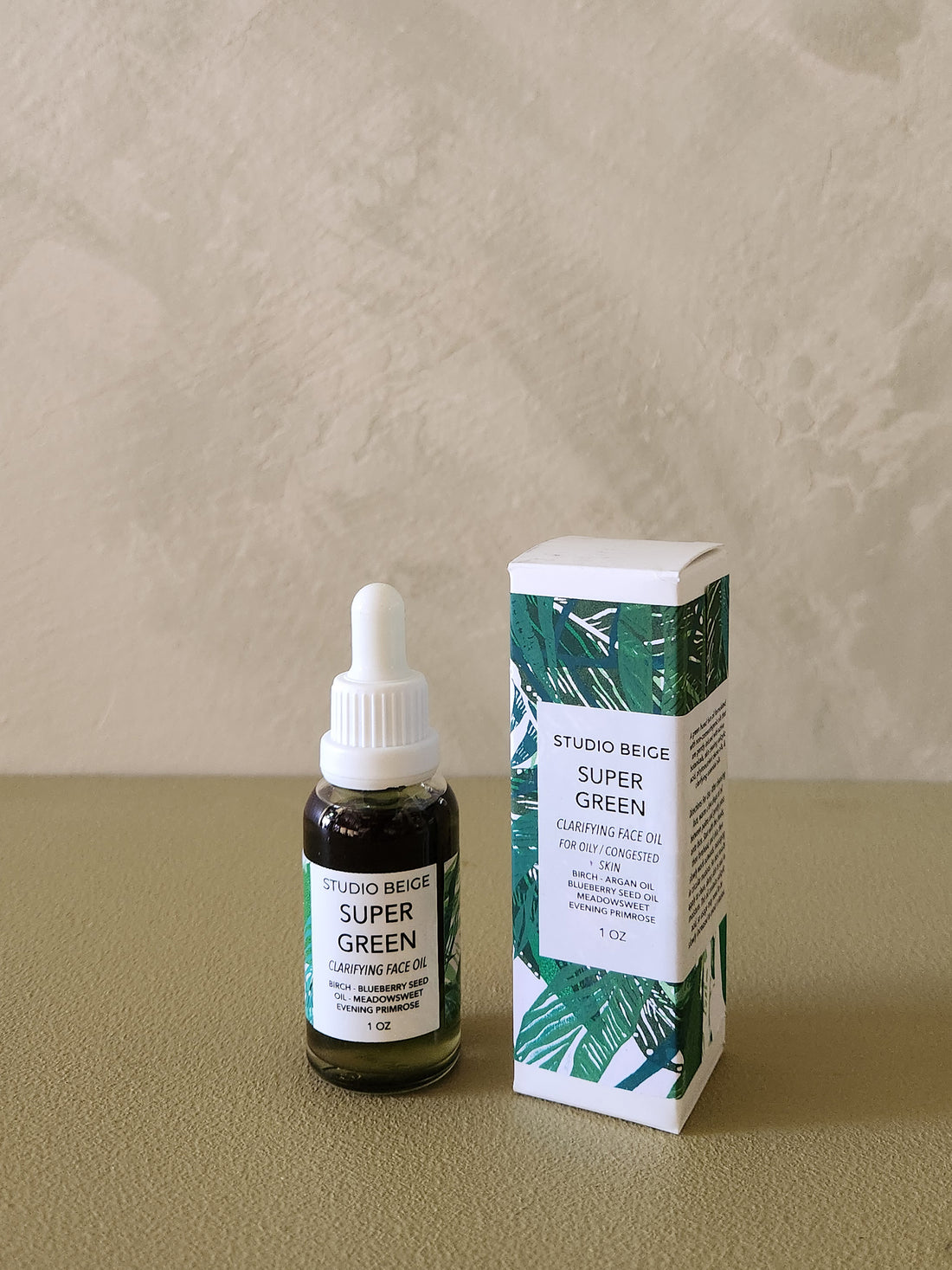 Bottle and box of Studio Beige Super Green face oil on a beige surface with a neutral background