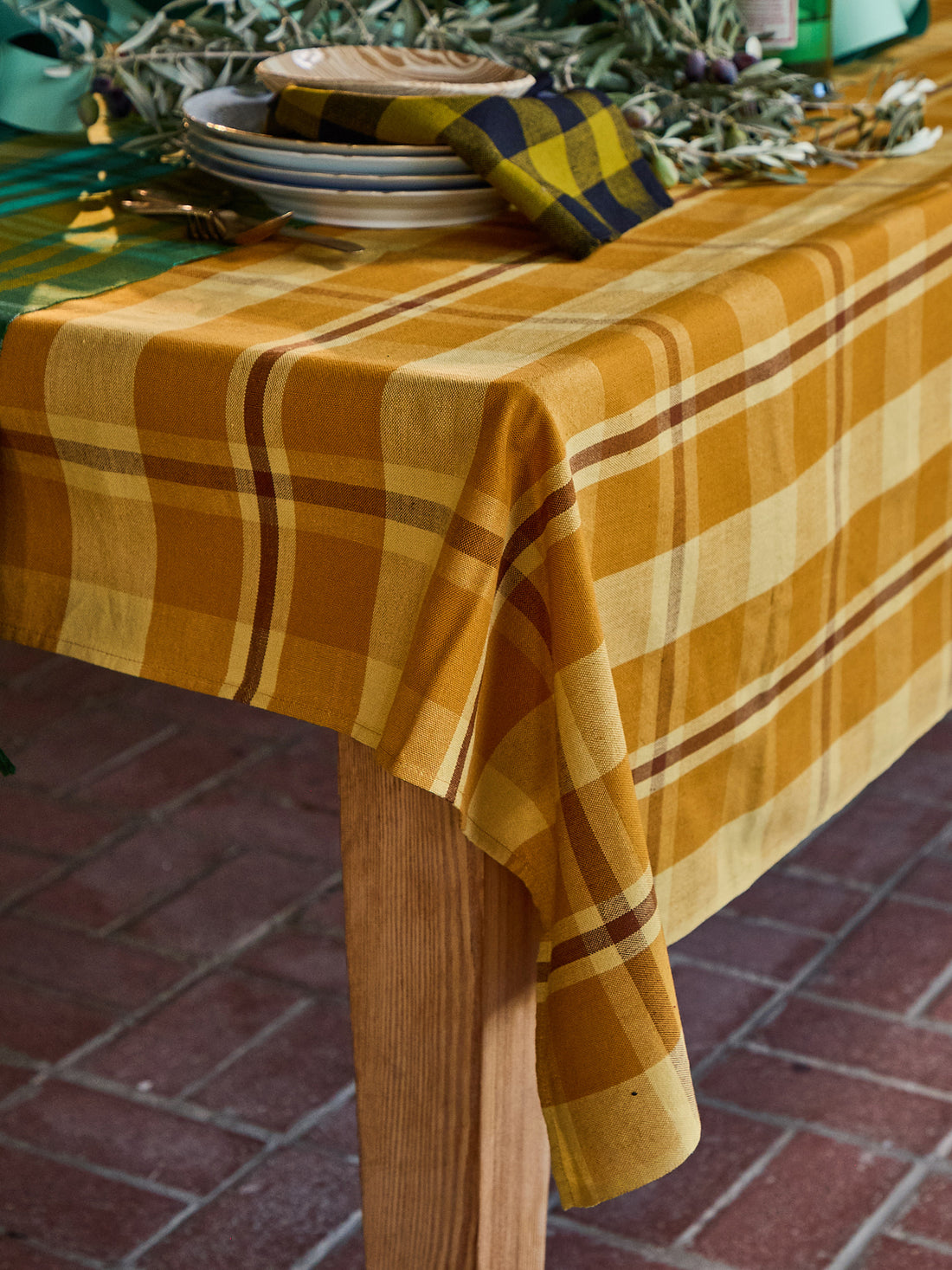 A mustard yellow and cream plaid tablecloth draped over a wooden table, styled with plates, a matching napkin, and greenery on a brick patio.