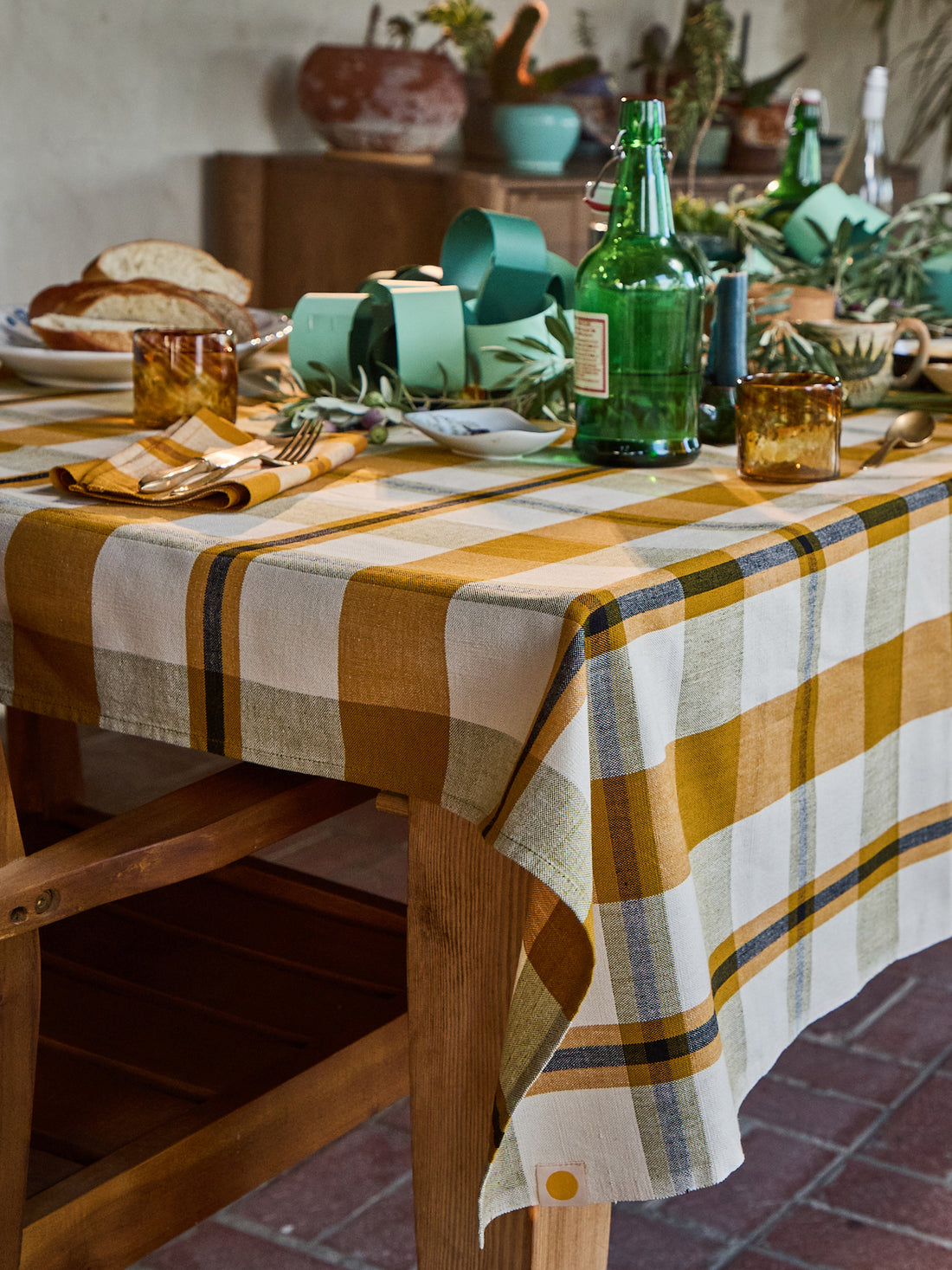 A rustic wooden dining table set with a yellow, white, and blue plaid tablecloth, amber glass tumblers, green glass bottles, and fresh herbs in a warm, styled dining room setting.