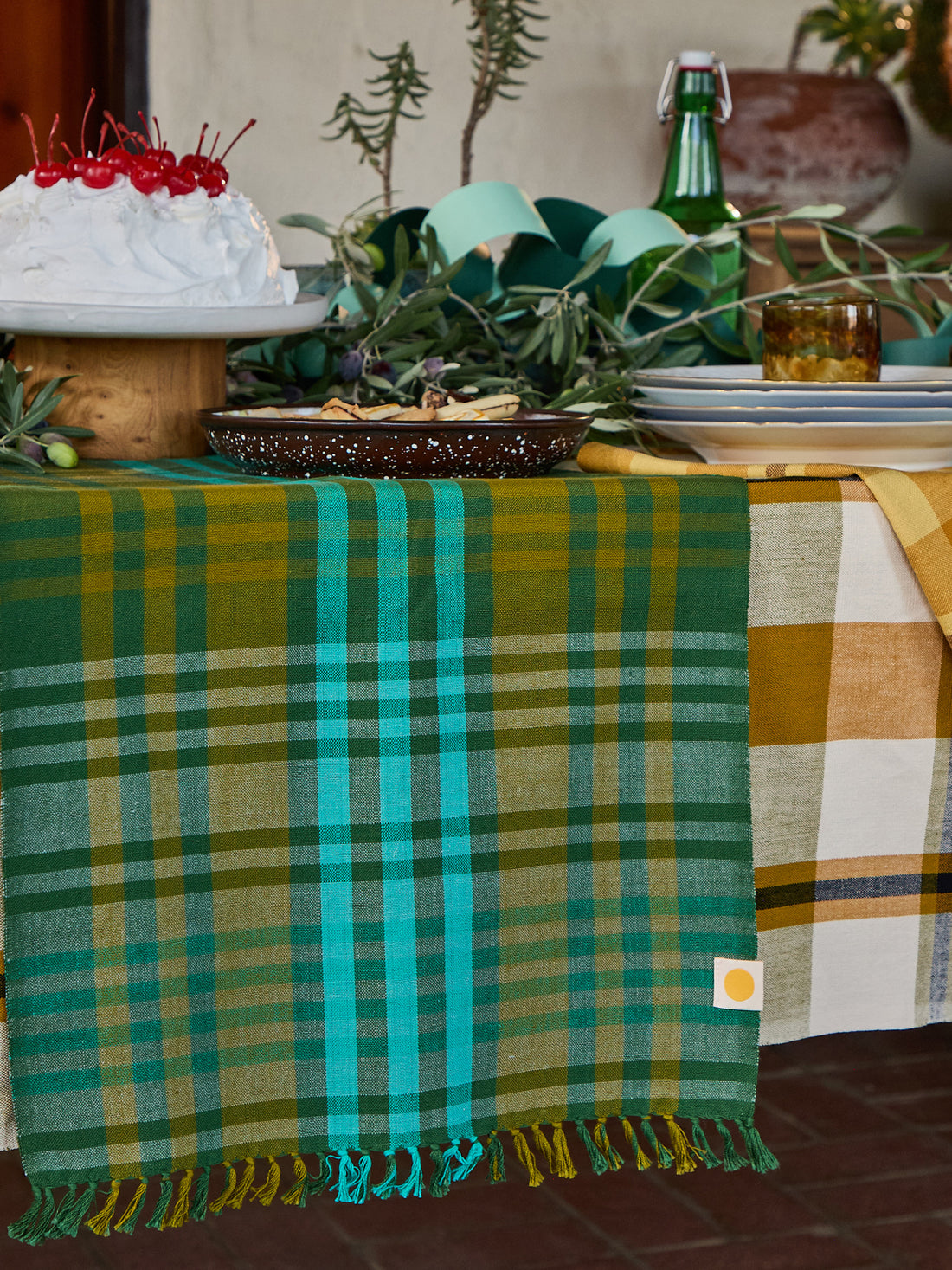 A green and teal plaid table runner with fringe is draped over a table, alongside a yellow-and-white plaid cloth. The scene is styled with dishes, a cake, greenery, and a bottle in a rustic indoor setting.
