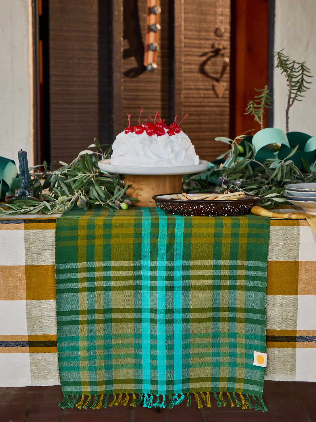 A green and teal plaid table runner with fringe is draped over a table decorated with a white cake topped with red cherries, greenery, and a bowl, set against a rustic wooden door background.