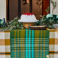 A green and teal plaid table runner with fringe is draped over a table decorated with a white cake topped with red cherries, greenery, and a bowl, set against a rustic wooden door background.
