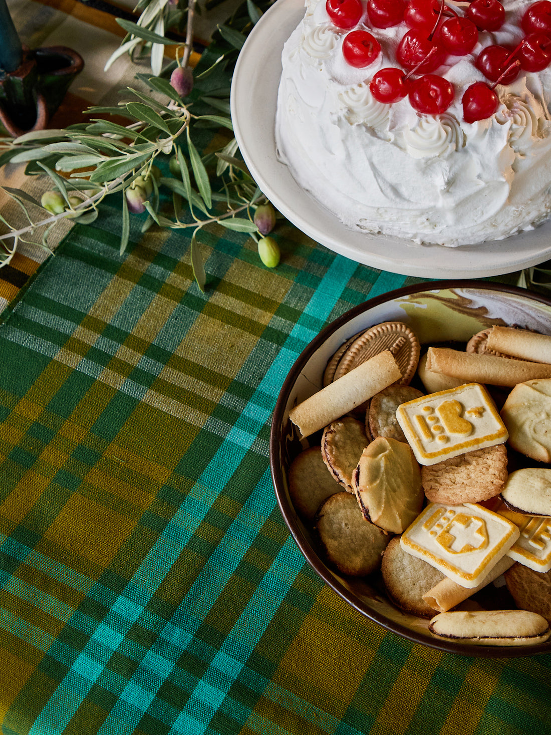 A styled tabletop scene with a plaid green and yellow tablecloth, a bowl of assorted cookies and biscuits, and a white whipped-cream cake topped with red cherries in the upper right.