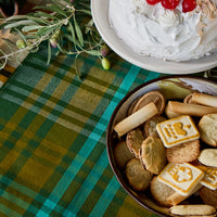 A styled tabletop scene with a plaid green and yellow tablecloth, a bowl of assorted cookies and biscuits, and a white whipped-cream cake topped with red cherries in the upper right.