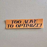 An orange rectangular bumper sticker with the text 'TOO ALIVE TO OPTIMIZE!' in black lettering, shown against a textured gray wall.