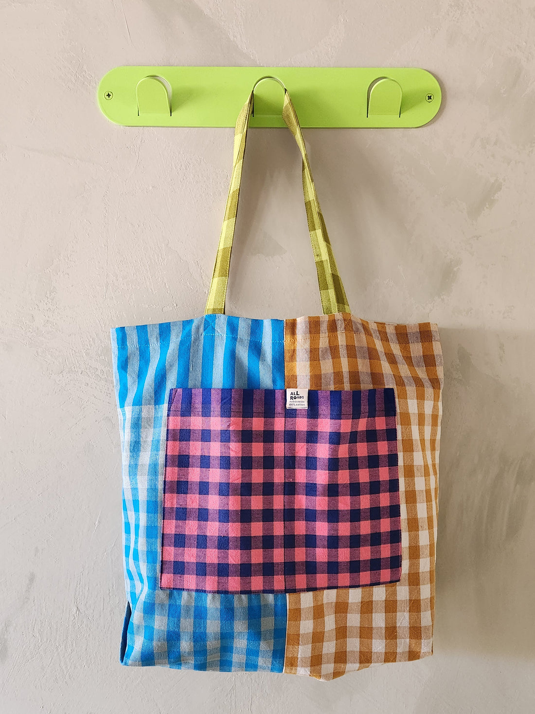 Colorful checkered tote bag with a pink pocket hanging on a green hook against a beige wall.