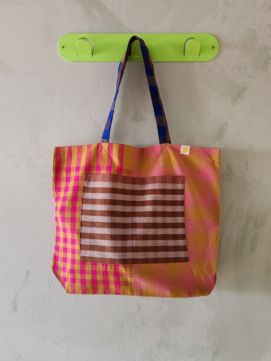 Colorful checkered bag with a striped pocket hanging on a green hook against a plain wall.
