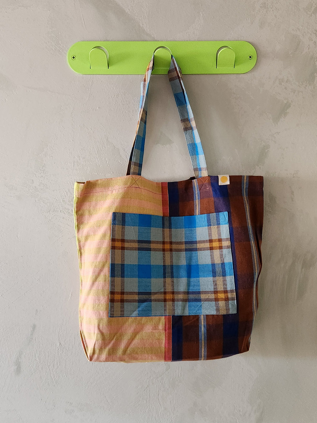 Colorful plaid tote bag hanging on a green hook against a beige wall.