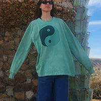 Person wearing a teal sweater with a yin-yang symbol in front of a stone wall and cactus.