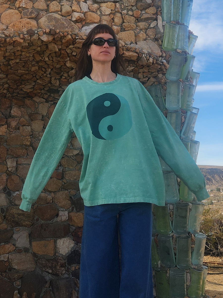 Person wearing a teal sweater with a yin-yang symbol in front of a stone wall and cactus.